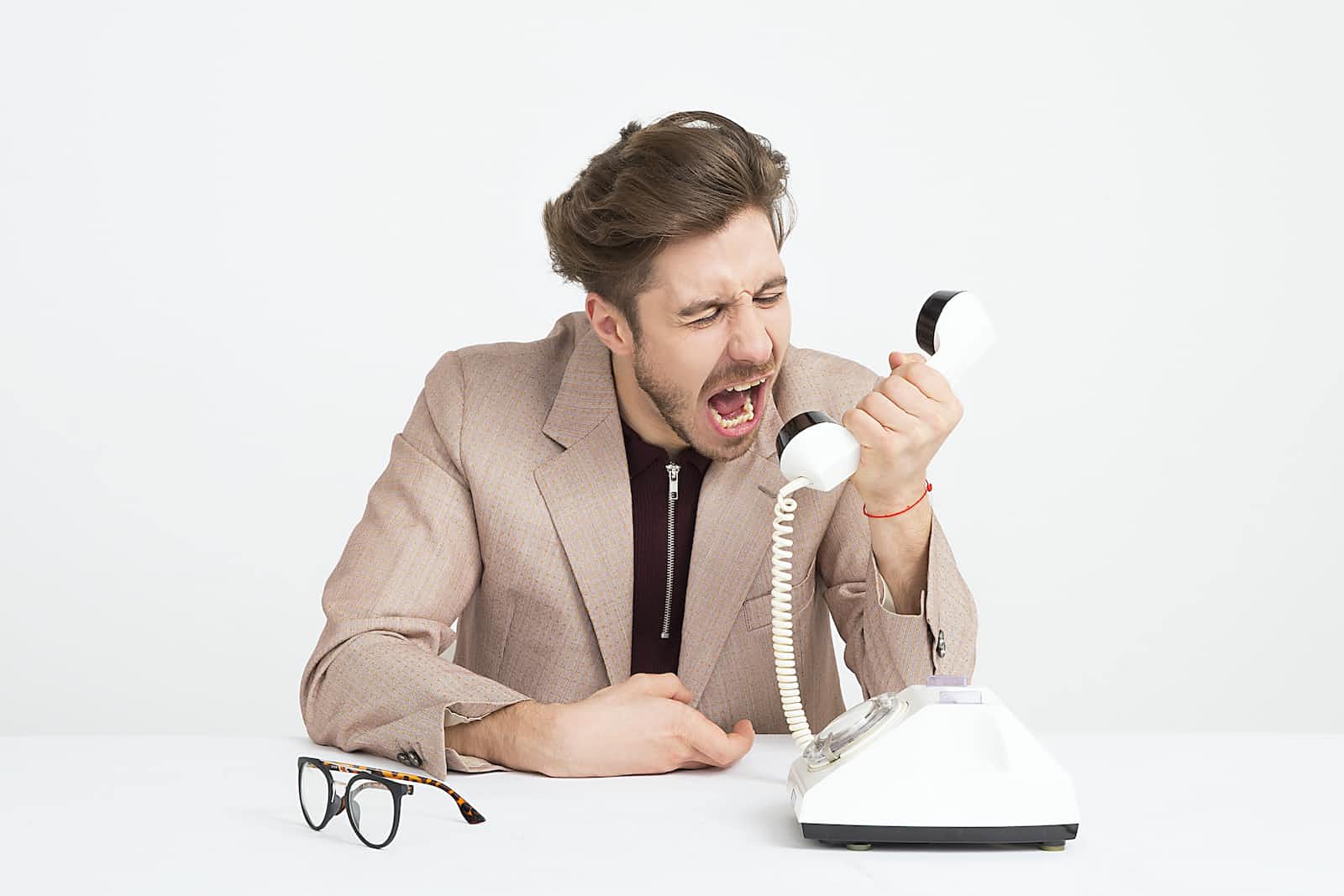 A man with his glasses removed, wearing a suit, angrily yelling at a telephone receiver